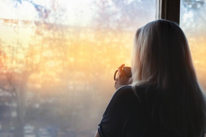 Woman standing by window with a cup looking out on a sunny day